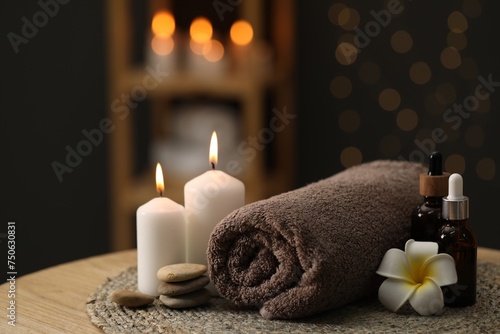 Spa composition. Rolled towel, cosmetic products, stones, burning candles and plumeria flower on table indoors, closeup. Space for text