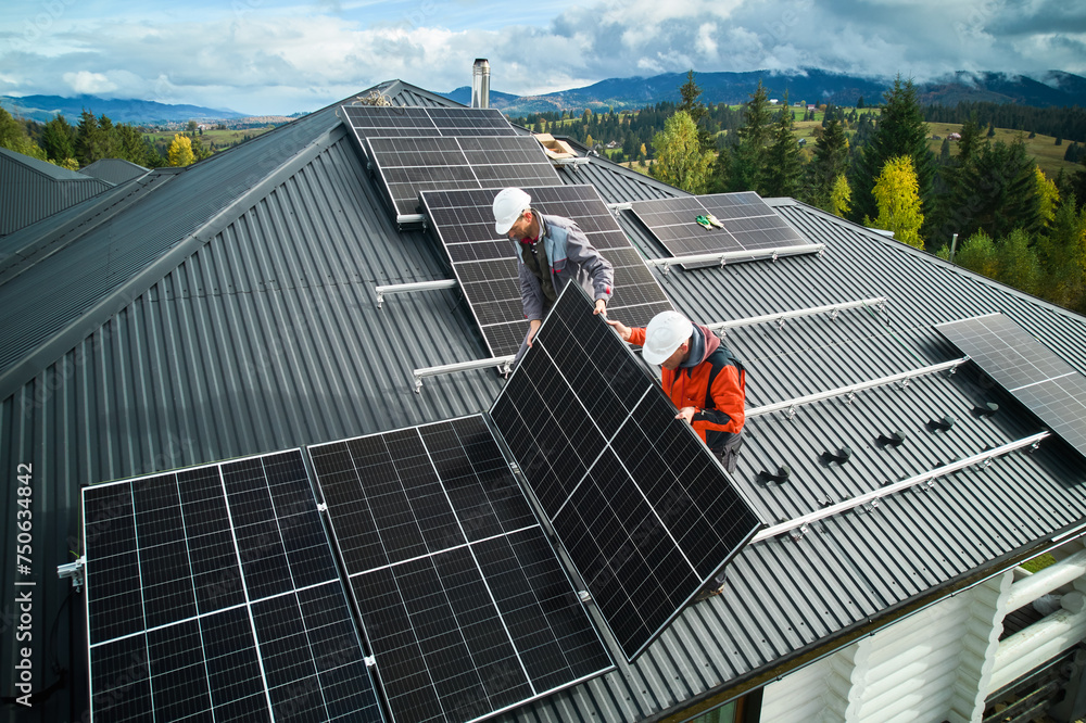 Roofers building photovoltaic solar module station on roof of house. Men electricians in helmets installing solar panel system outdoors. Concept of alternative and renewable energy. Aerial view. - obrazy, fototapety, plakaty 