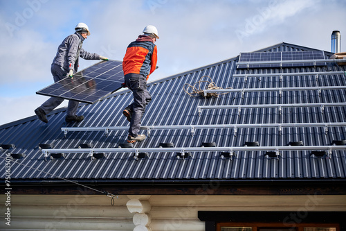 Roofers building solar panel system on roof of house. Men workers in helmets carrying photovoltaic solar module outdoors. Concept of alternative and renewable energy.