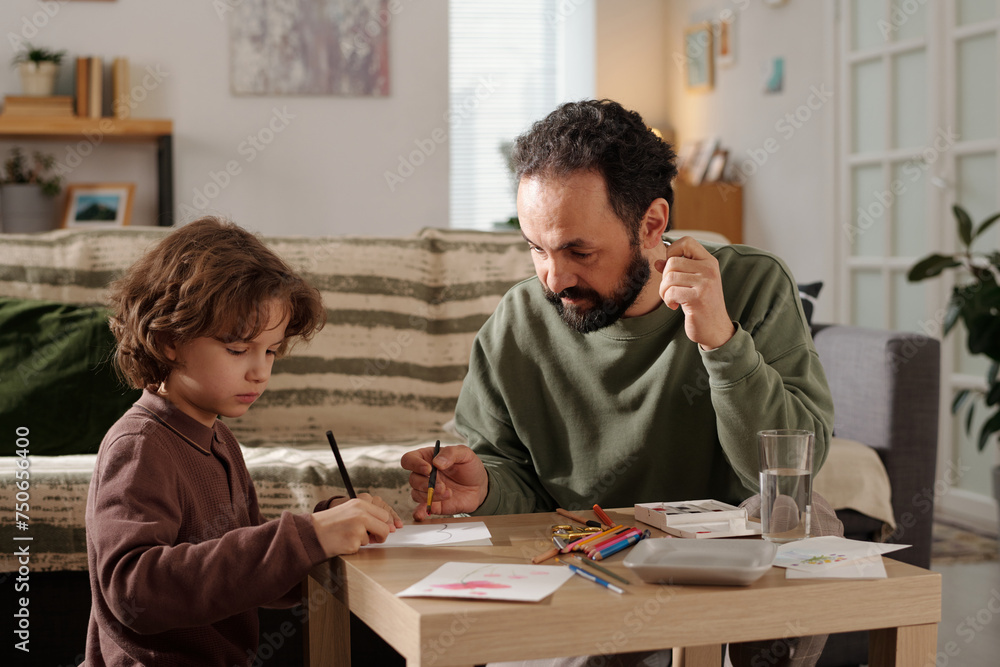 Young man with paintbrush in hand looking at his son drawing with crayons while both preparing handmade postcard for mother day Young man with paintbrush in hand looking at his son drawing with crayons while both preparing handmade postcard for mother day
