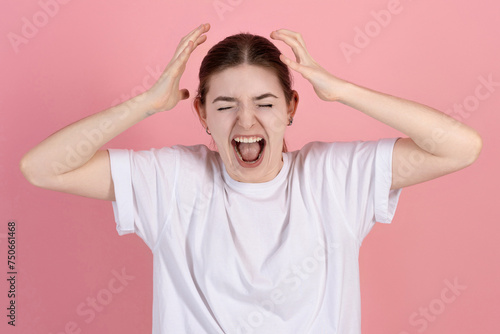 Angry attractive caucasian young brunette woman in casual white t-shirt with eyes closed yells loudly isolated on pink studio background.