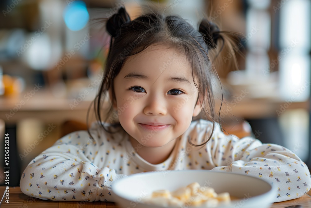 Cheerful young girl with playful pigtails smiles warmly while sitting in front of a breakfast bowl at a home setting. Cheerful young girl with playful pigtails smiles warmly while sitting in front of a breakfast bowl at a home setting.