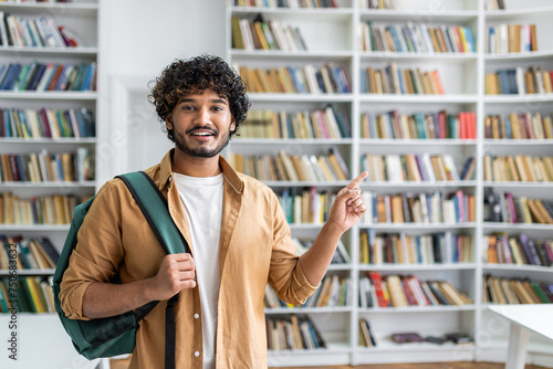 A happy young man with curly hair and a casual outfit is standing in a library. He is carrying a backpack on one shoulder and pointing at something out of frame with a smile on his face.