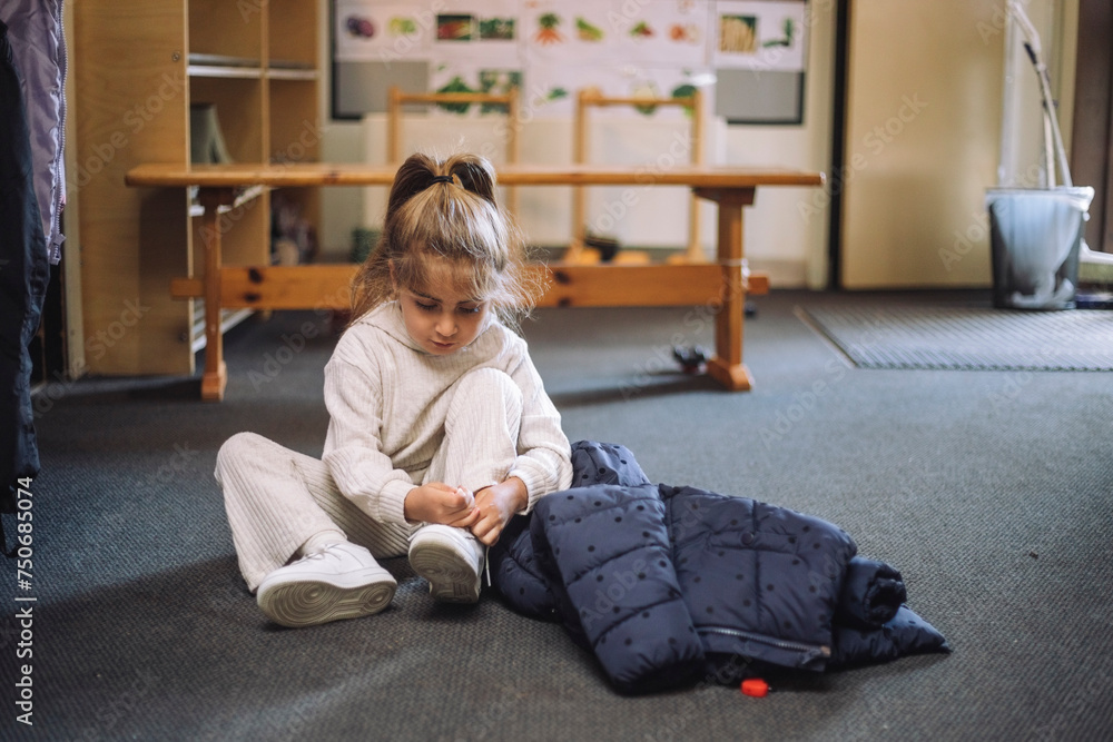 Girl tying shoelace while sitting with jacket on floor in classroom at kindergarten Girl tying shoelace while sitting with jacket on floor in classroom at kindergarten