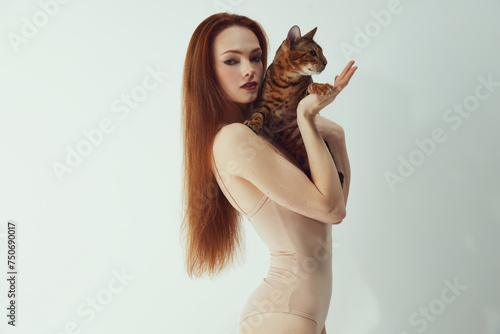 Quadro em tela Portrait of elegant, beautiful, young redhead woman in beige bodysuit holding Bengal cat against white studio background