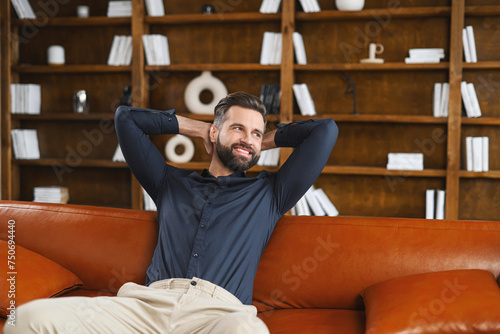 A man is lounging comfortably on a couch, with a smile on his face and his hands behind his head. He is wearing a smart casual shirt, looking happy and relaxed, ook shelves on the background photo