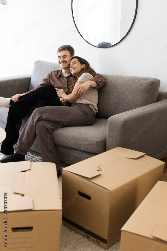 Happy couple embracing while sitting on sofa by cardboard boxes at home Happy couple embracing while sitting on sofa by cardboard boxes at home