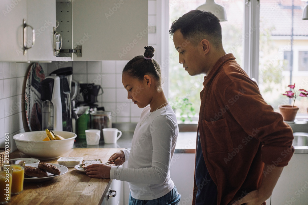 Side view of father looking at daughter preparing breakfast while standing in kitchen at home Side view of father looking at daughter preparing breakfast while standing in kitchen at home