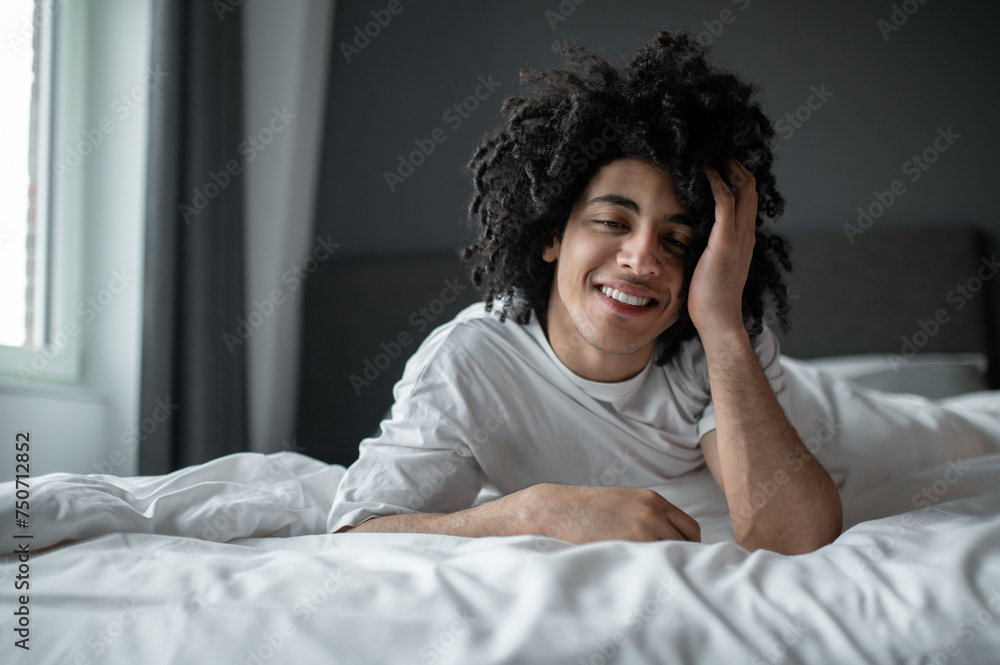 Young curly-haired man lying in bed and looking relaxed and sleepy Young curly-haired man lying in bed and looking relaxed and sleepy