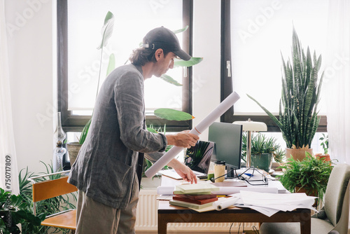 Side view of male architect holding rolled blueprint while standing at desk in home office
