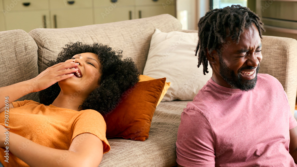Laughing Couple At Home With Woman Lying On Sofa And Man Sitting On Floor Laughing Couple At Home With Woman Lying On Sofa And Man Sitting On Floor
