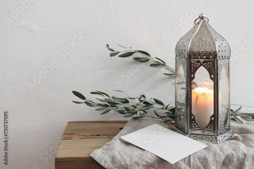 Ramadan Kareem holiday still life. Ornamental silver Moroccan lantern with olive tree branches. Blank greeting card, invitation mockup on wooden table, bench. Blurred white wall background.