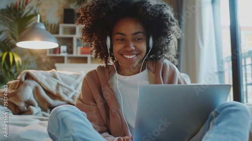 Beautiful young African American girl with headphones and laptop sitting with folded legs on the floor in a cozy room. Smiling black female student learning online. Remote education concept.