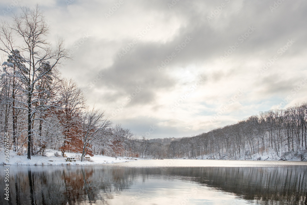 A wide angle view of a small lake in Indiana in the middle of winter on a snowy day with a mostly cloudy sky. The sun is bursting through the clouds and there are nice reflections on the water. A wide angle view of a small lake in Indiana in the middle of winter on a snowy day with a mostly cloudy sky. The sun is bursting through the clouds and there are nice reflections on the water.