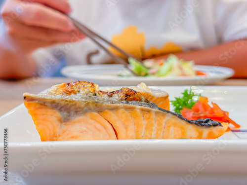 Selective focus of grilled salmon steak on white plate, Japanese food style with blurry women hand and chopsticks