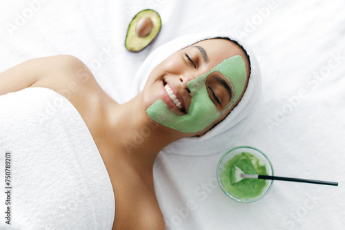 Skin care concept, facial cleansing. Top view of a gorgeous young woman of brazilian or hispanic ethnicity, with eyes closed lying in a beauty salon with a green clay mask on her face, smiling