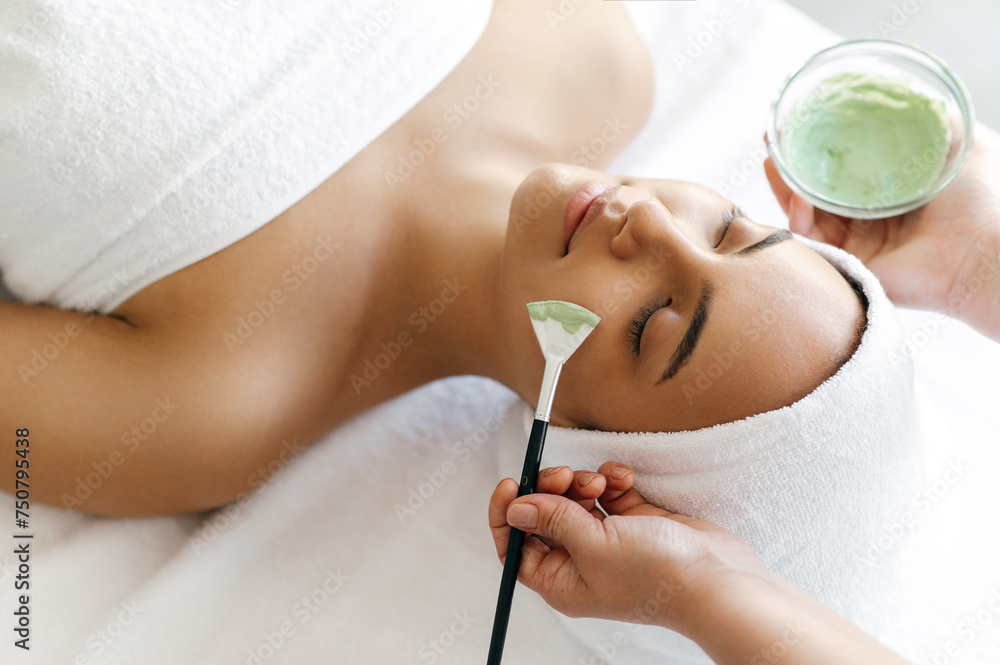 Naklejka premium Facial treatments, wrinkle prevention, facial cleansing. Top view of beauty procedure, therapist applying green face mask on the face of a beautiful brazilian or hispanic young woman