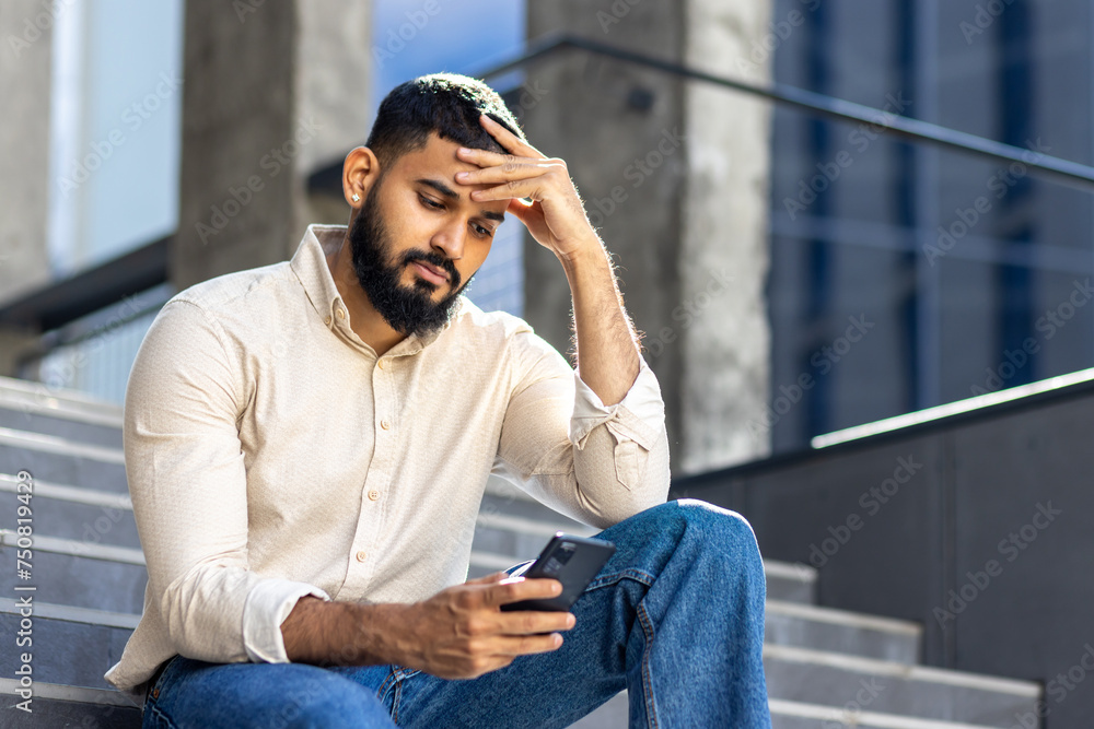Photo of a sad young Muslim man sitting near a building on the stairs, holding his head and looking worriedly at the mobile phone screen Photo of a sad young Muslim man sitting near a building on the stairs, holding his head and looking worriedly at the mobile phone screen