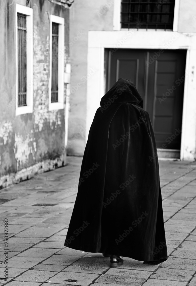 hooded anonymous person walking wearing an old black tabard as a cloak on the narrow alley of the city hooded anonymous person walking wearing an old black tabard as a cloak on the narrow alley of the city