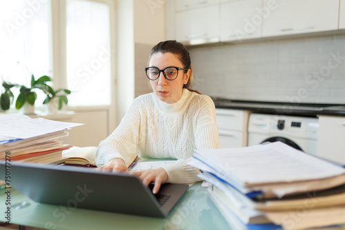 woman with glasses managing business affairs on her laptop, amidst scattered documents in her home