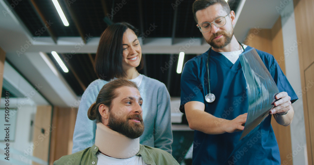 Obraz premium Portrait view of cute young nurse helping patient get around the hospital in wheelchair. Attractive male Caucasian doctor examining x-ray and discussing injury. Modern clinic concept.