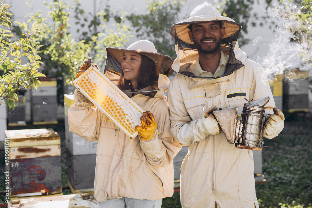 Couple of happy smiling beekeepers working with beekeeping tools near beehive at bee farm Couple of happy smiling beekeepers working with beekeeping tools near beehive at bee farm