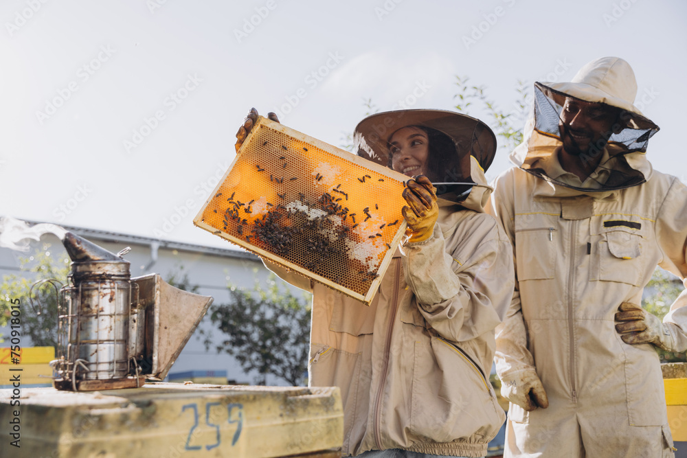 Couple of happy smiling beekeepers working with beekeeping tools near beehive at bee farm - obrazy, fototapety, plakaty 