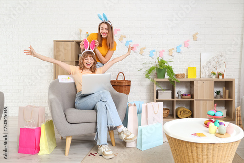 Happy young women in bunny ears headbands with laptop and paper bags for Easter at home. Online shopping concept