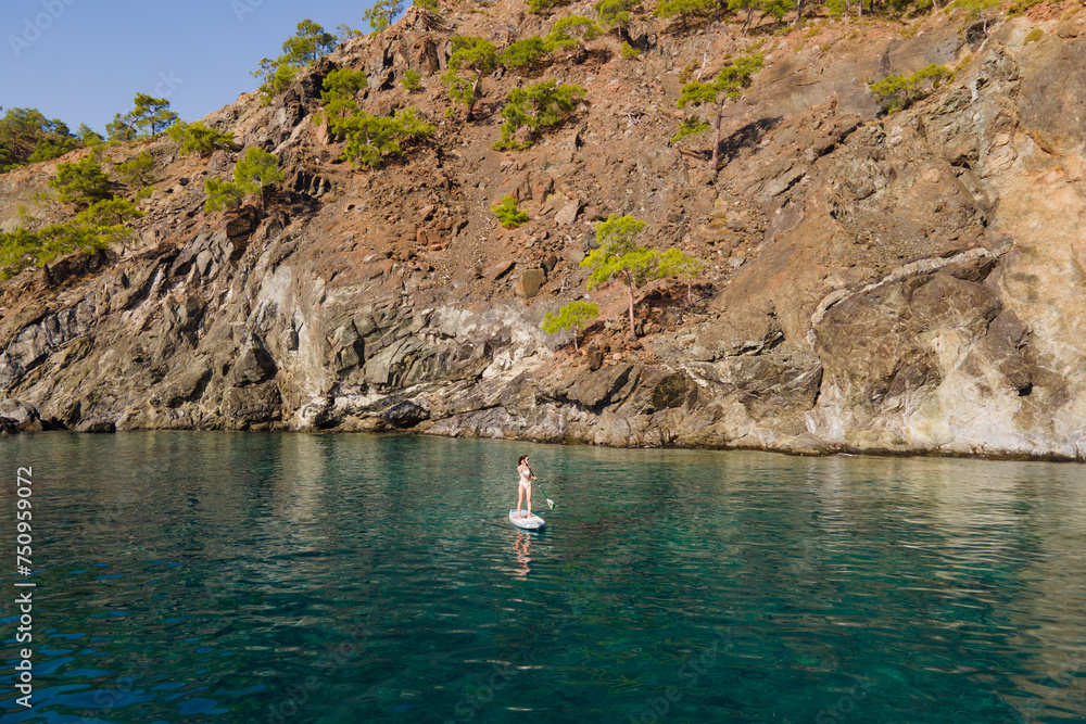 Aerial view of woman paddling a stand-up paddleboard or SUP board on a calm sea along rocky shore