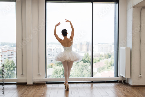 Back view of focused young Caucasian ballerina dressed in white tutu costume. Beautiful female ballet dancer makes pirouette at ballet studio in front of big panoramic window background. photo