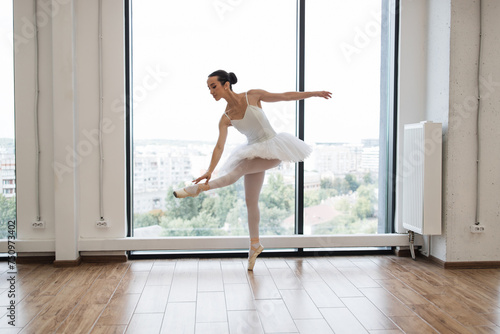 Caucasian ballerina posing in bright class studio with panoramic windows. Young graceful ballet dancer rehearsing performance in white spacious hall.