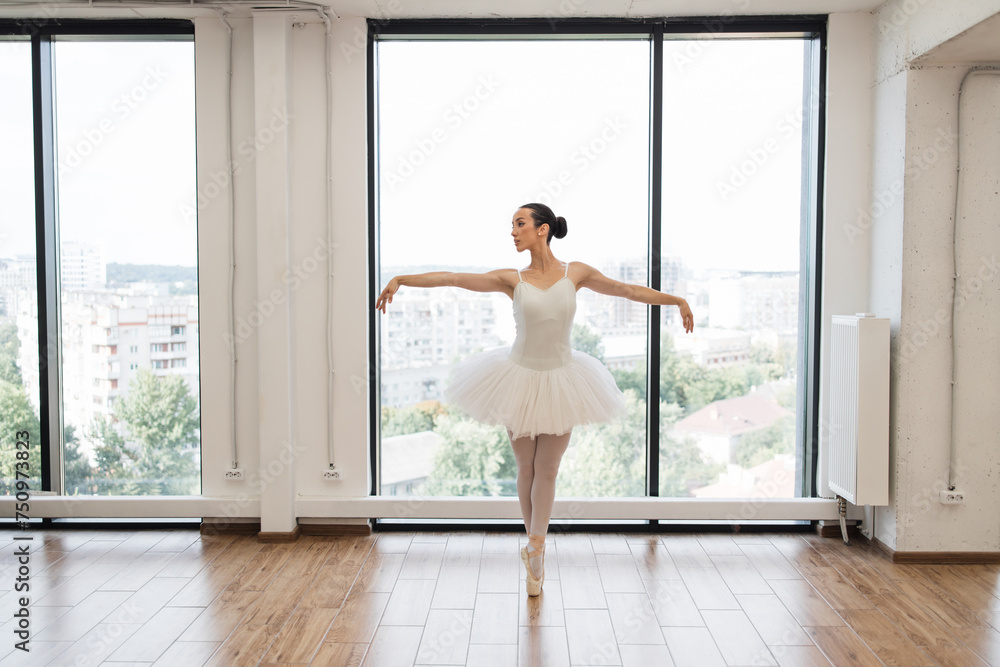 Young graceful ballet dancer rehearsing performance in white spacious hall. Caucasian ballerina posing in bright class studio with panoramic windows. Young graceful ballet dancer rehearsing performance in white spacious hall. Caucasian ballerina posing in bright class studio with panoramic windows.