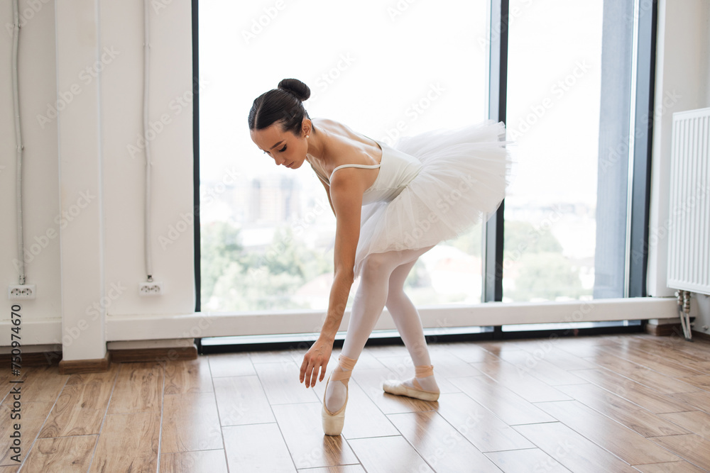 Young graceful ballet dancer rehearsing performance in white hall room. Caucasian ballerina stretching to feet while standing in dance studio with large windows. Young graceful ballet dancer rehearsing performance in white hall room. Caucasian ballerina stretching to feet while standing in dance studio with large windows.