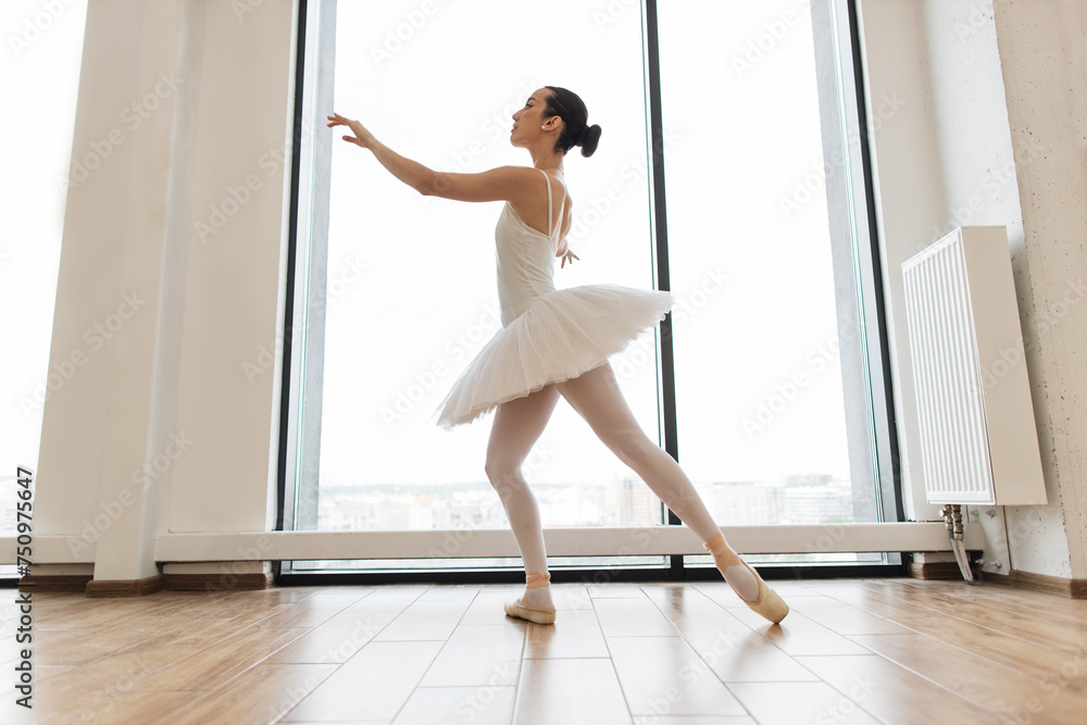 Beautiful ballerina practice ballet positions in white skirt near large window in white hall. Young ballet dancer - Harmonious pretty woman with tutu posing in studio - Contemporary dance performer.