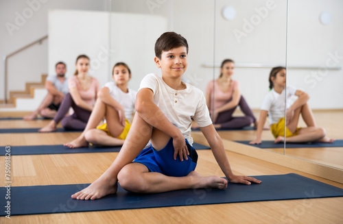 Smiling flexible teen boy sitting on mat in twisting asana Matsyendrasana during yoga class with sportive family in fitness studio