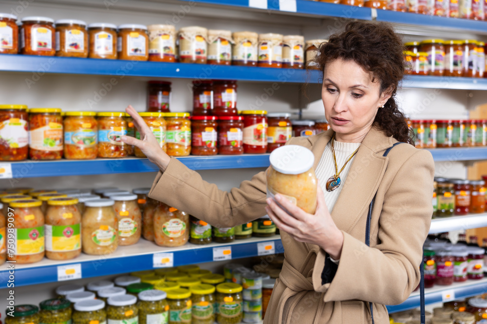 Woman in store of imported Russian goods chooses jar with canned fermented cabbage. Female buyer is looking for traditional vegetable national Russian snacks salad, imported products in store