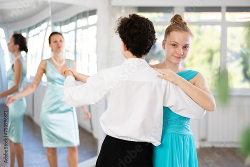 Motivated adolescent ballroom dancers, girl and boy in performance outfit practicing elegant dance moves in pair in bright studio, with female coach supervising in background
