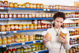 Female shopper selects glass jar of sauerkraut at a grocery store. Woman shopper reading expiration date on product label