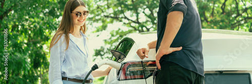 Focused EV car recharging electricity for battery on blurred background of lovey couple ruing their road trip travel by eco friendly electric car in national park and greenery forest on holiday. Exalt photo