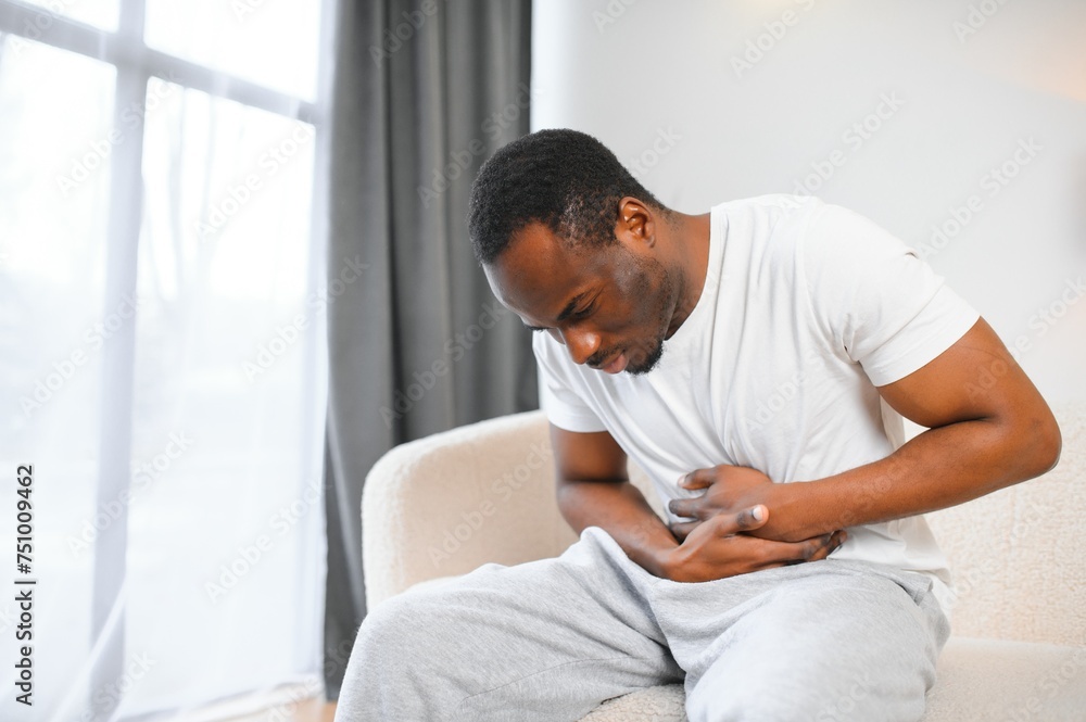 African american guy having stomach ache after eating touching aching stomach suffering from pain sitting on sofa at home. African american guy having stomach ache after eating touching aching stomach suffering from pain sitting on sofa at home.