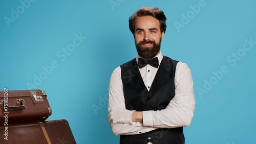 Professional hotel concierge posing on camera against blue background, preparing to carry luggage and help travellers. Elegant skilled employee working as bellboy, tourism industry concept. photo