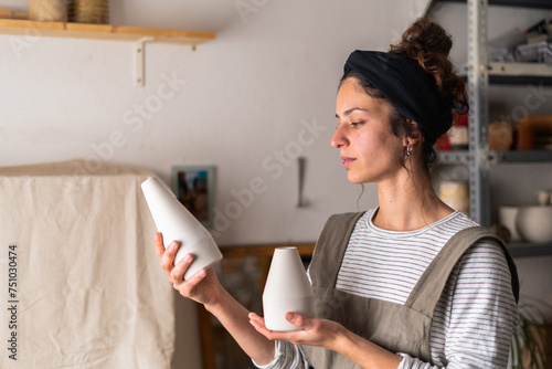 A focused female ceramist examines the shape and size of two unfinished ceramic vases in her workshop