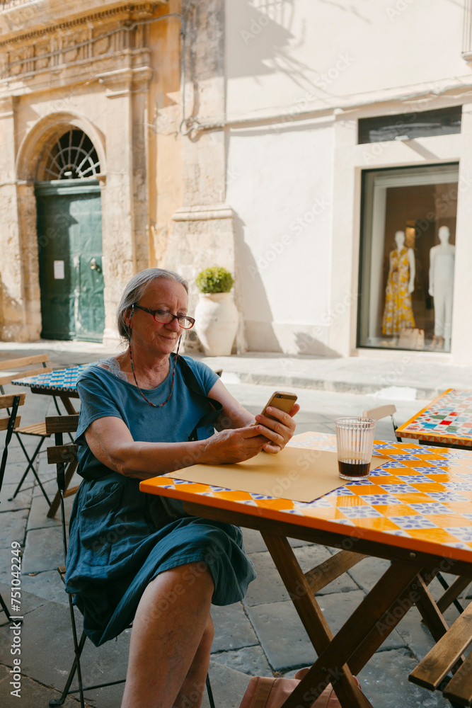 © Jimena Roquero/Stocksy - Senior woman sitting on a colorful outdoor cafe checking her phone