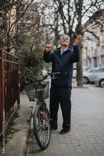 An elderly man in casual attire is inspecting tree branches with curiosity near his parked bicycle in an urban setting, capturing a moment of leisure.