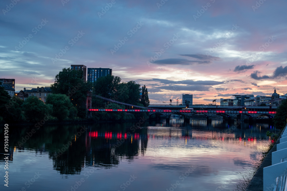 Experience the poetic allure of twilight with this enchanting photo featuring a bridge adorned with radiant red lights gracefully spanning over a tranquil river. 