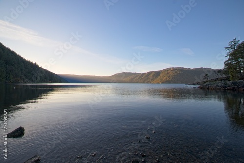 The tranquil waters of a calm lake mirror the fading hues of the day, set against a backdrop of majestic mountains in the distance.