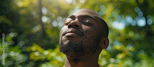 A black man is standing with his eyes closed, facing upwards in a park. He seems to be enjoying the refreshing outdoor environment.