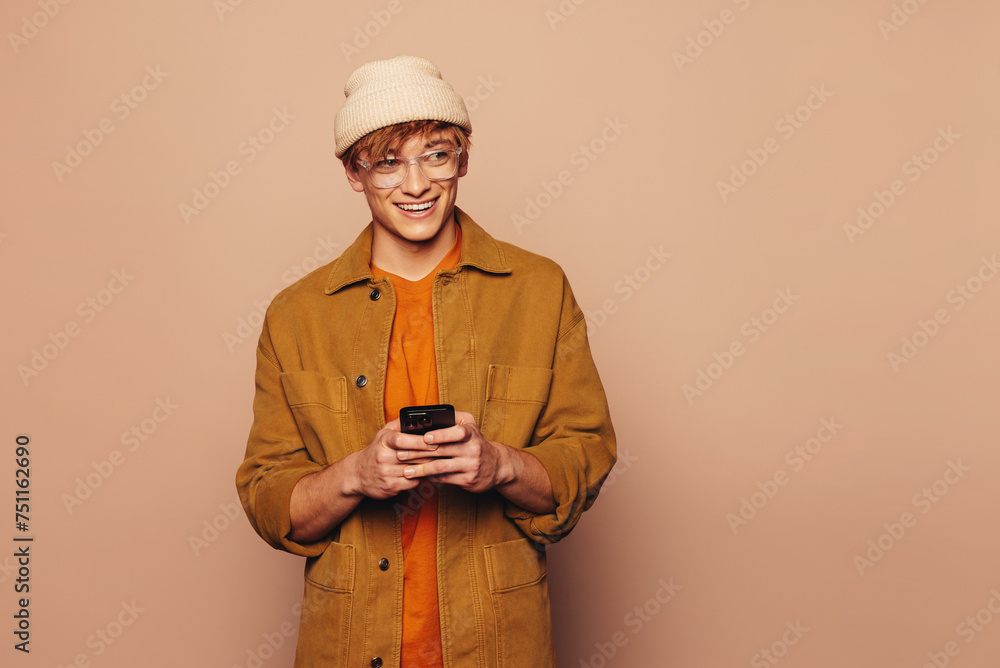Cheerful man using smartphone while standing in front of vibrant peach background - obrazy, fototapety, plakaty 