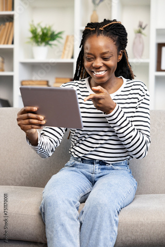 An upbeat Afro American woman with a bright smile engages in a video call using her tablet while comfortably seated on her couch, embodying a cozy, relaxed lifestyle.