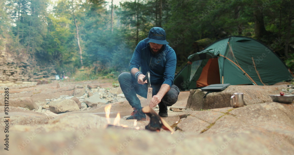 a male tourist chops firewood with a small ax near a campfire in the forest. the traveler prepares wood for the fire. man in the wild. outdoor hiker, wilderness survival, campfire a male tourist chops firewood with a small ax near a campfire in the forest. the traveler prepares wood for the fire. man in the wild. outdoor hiker, wilderness survival, campfire